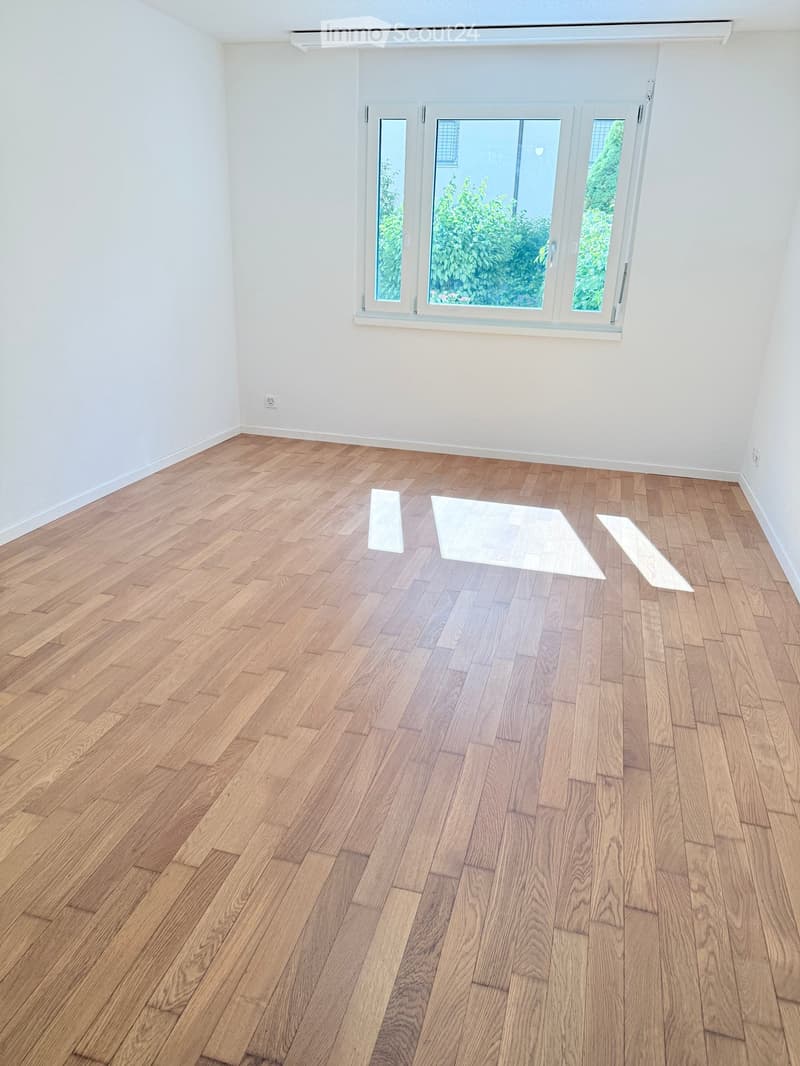 Empty room with light wooden herringbone flooring and white walls, featuring large windows with white frames and a view of greenery outside.