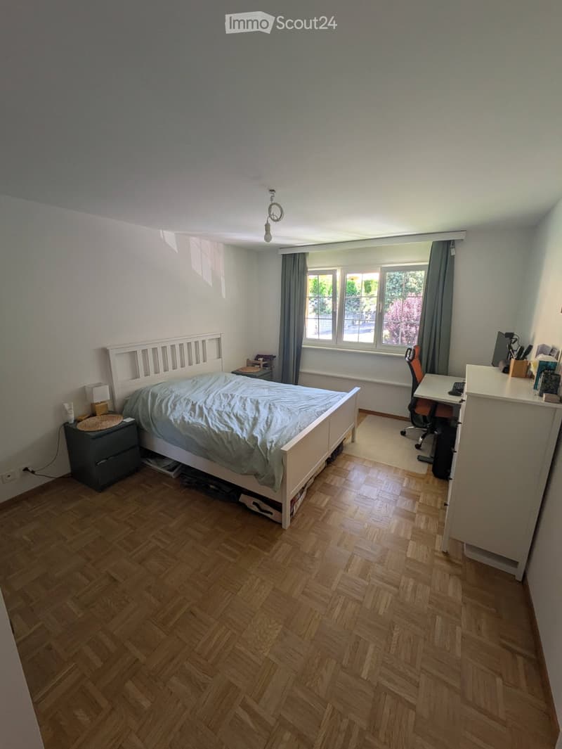 Bedroom with wooden herringbone flooring, a white bed with a blue duvet, a nightstand with a lamp, a desk with a chair, and large windows with grey curtains.