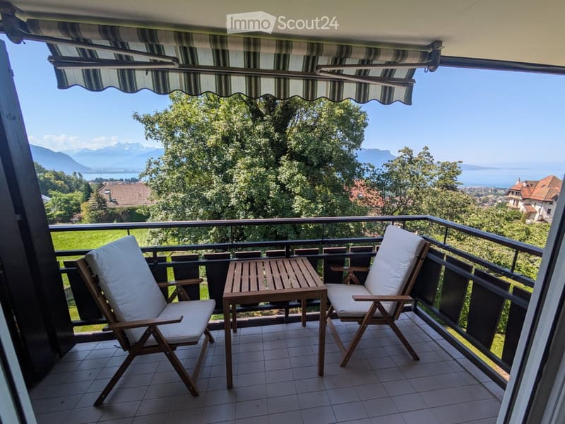 Balcony with two wooden chairs and a small table, overlooking a scenic view of mountains and a lake.