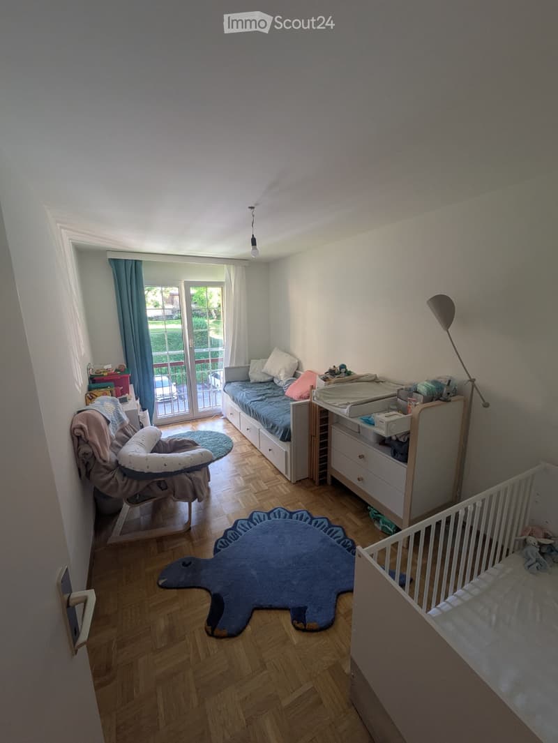A well-lit children's bedroom with a baby bed, changing table, rocking chair, and a large dinosaur-shaped rug.