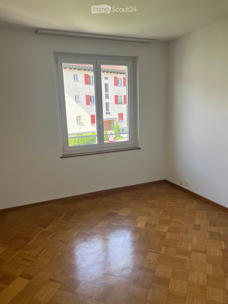 Empty room with wooden parquet flooring, white walls, and a large window with a view of a neighboring building.