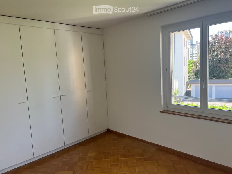 Empty room with wooden flooring, white built-in wardrobes, and a large window offering a view of the city and greenery.