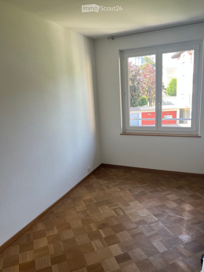 Empty room with wooden parquet flooring and white walls, featuring a large window with a view of trees and buildings.