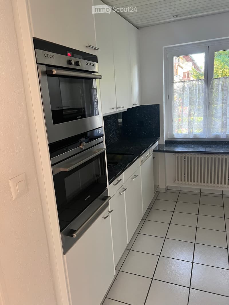 Modern kitchen with white cabinets, black countertop, and a built-in oven. The space features tiled flooring and a window with sheer curtains.