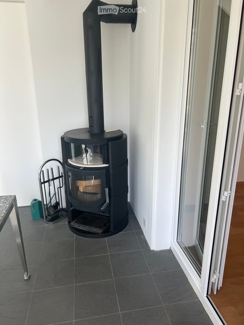 Modern black wood-burning stove with glass front, firewood rack, and a small table near the window.