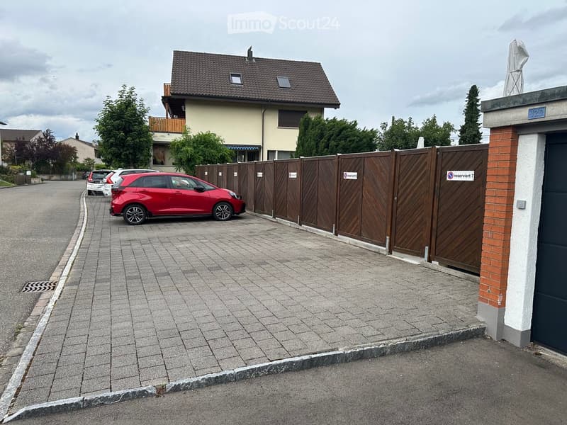 A two-story residential building with a tiled roof, surrounded by a wooden fence and a paved driveway with several parked cars.