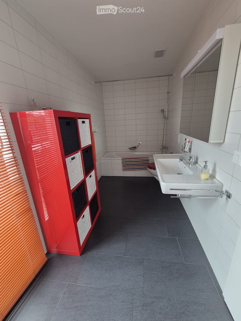 Modern bathroom with white tiles, a red storage cabinet, a white sink with a mirror, a bathtub, and a toilet. Features include a shower area and a soap dispenser on the sink counter.