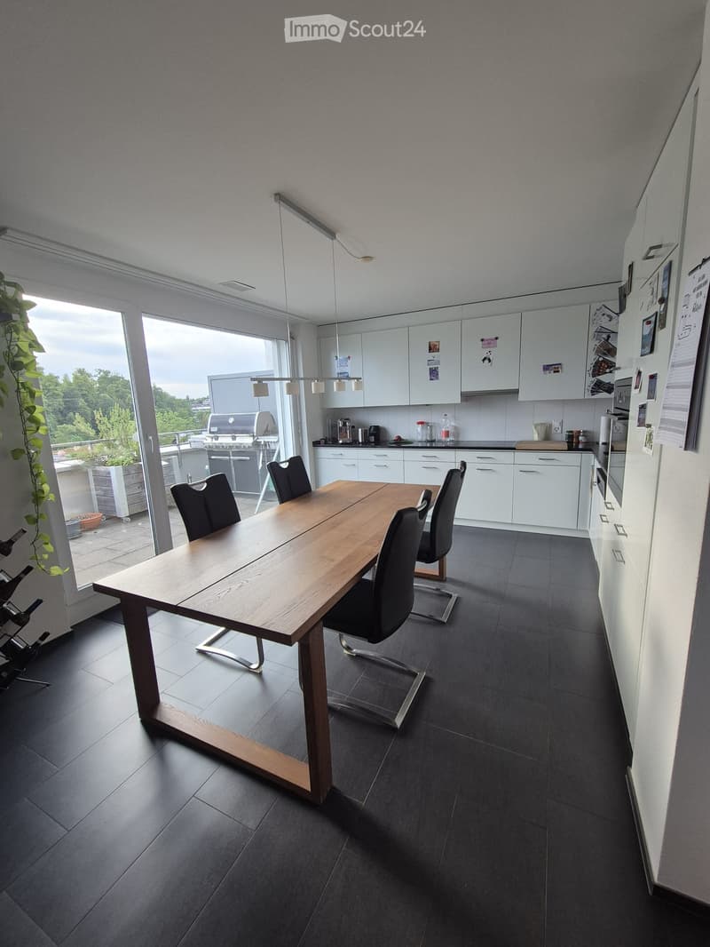 Modern kitchen with a large wooden dining table and four black chairs, white cabinets, and a refrigerator. The room features dark tiled flooring and large windows with a view of the outdoor area.