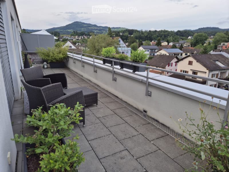 Balcony with wicker chairs, small table, potted plants, and scenic mountain view.