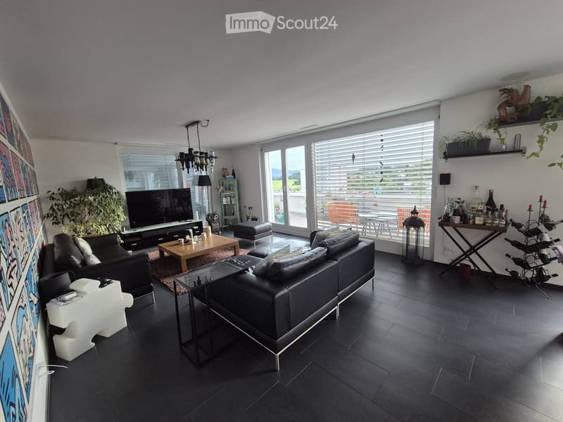 Modern living room with dark tiled flooring, large windows, black leather sofa, coffee table, ottoman, TV, side table, wine rack, and decorative plants.