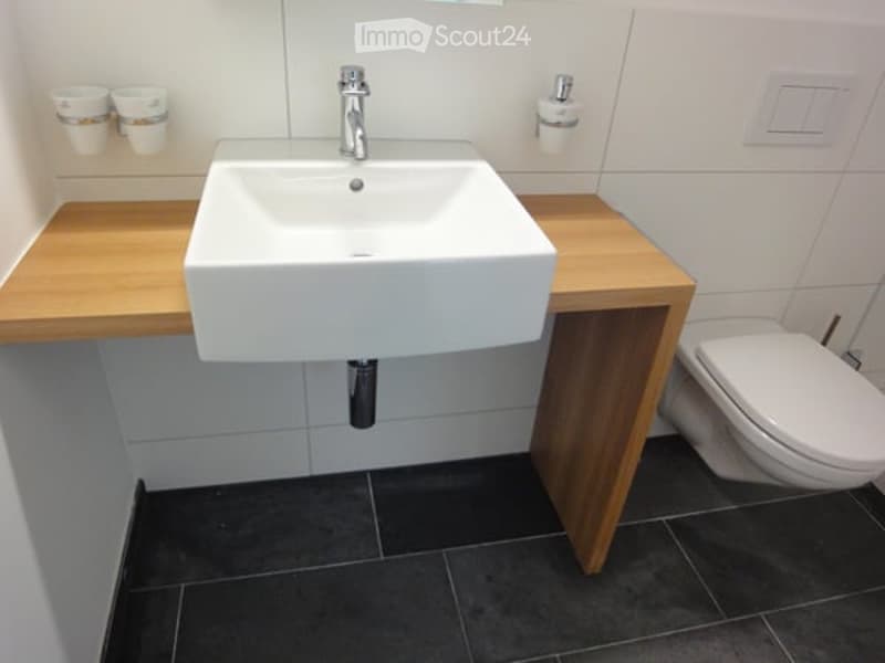 Modern bathroom with a white sink, chrome faucet, and a toilet. The sink sits on a light wooden vanity, and the floor is covered with dark tiles.