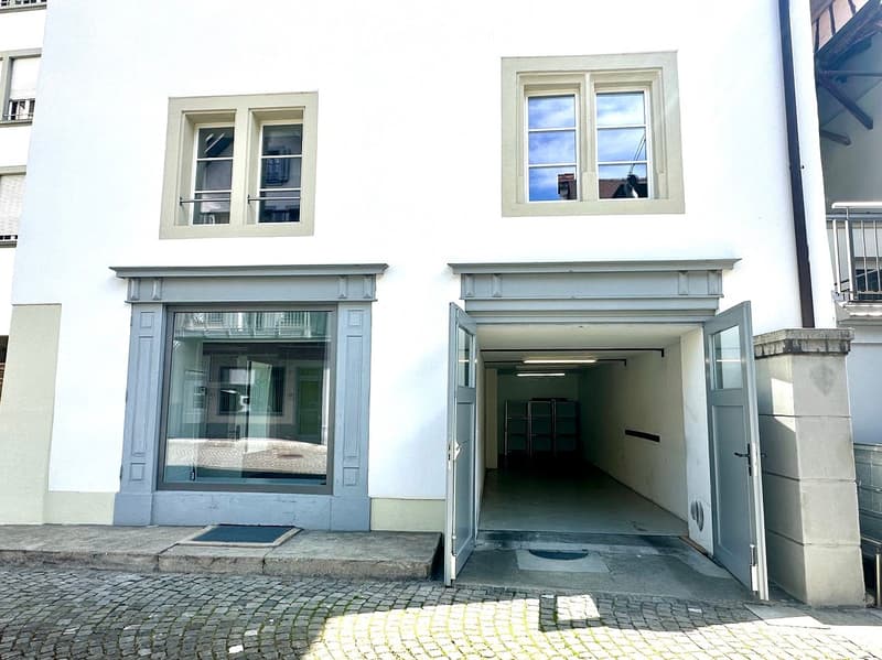 Two-story building with a garage entrance, large windows, and a cobblestone sidewalk.