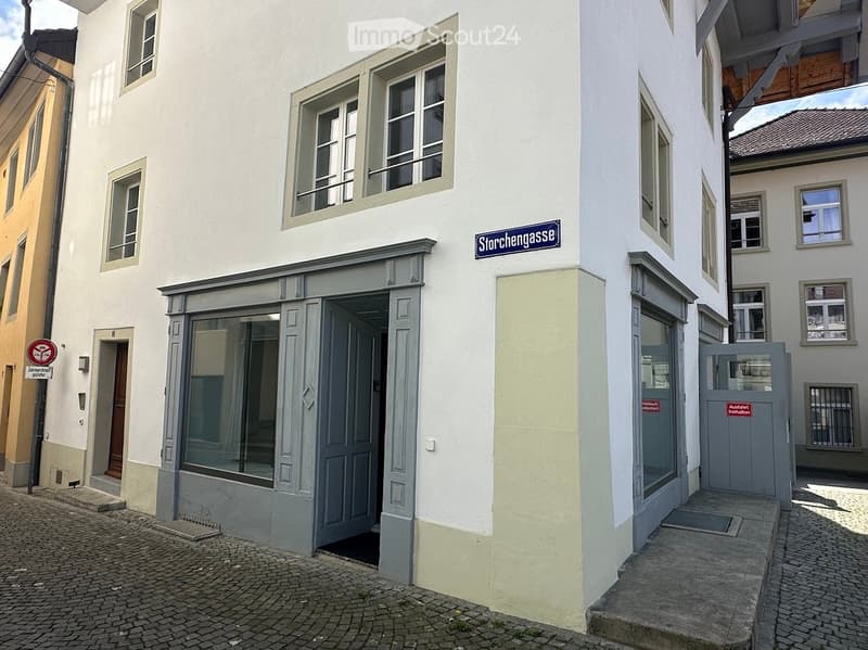Two-story building with white facade, multiple windows, and a storefront entrance with glass doors. The building is located on a cobblestone street with a street sign reading "Störchengasse" visible.