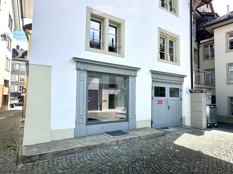 A three-story white building with a storefront on the ground floor, featuring large glass windows and a grey door. The upper floors have multiple windows with white frames, and the building is situated on a cobblestone street.