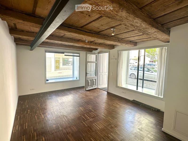 Empty room with wooden flooring, exposed wooden ceiling beams, large windows, and a door leading outside.