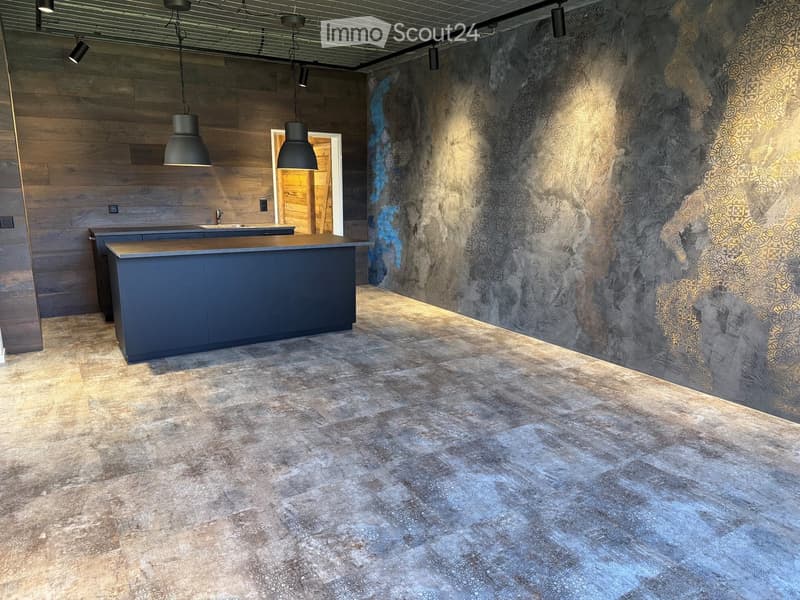 Modern kitchen with a dark grey island, wooden accents, and a textured wall design. The space features pendant lights and a polished concrete floor.