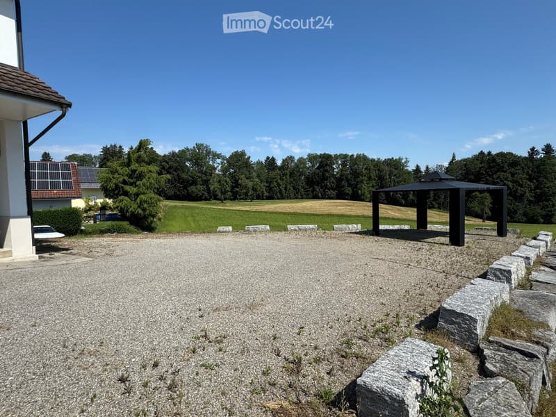 Gravel driveway leading to a house with a solar panel roof, surrounded by a grassy field and trees. A black gazebo is visible in the distance.