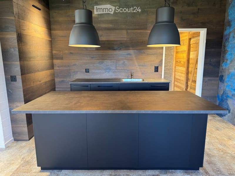 Modern kitchen with a large central island, dark grey cabinets, and a wooden countertop. The backsplash features a mix of light and dark wood panels, and the space is illuminated by two black pendant lights.