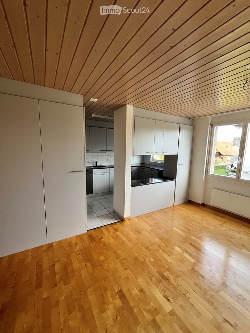 Empty room with wooden flooring, white walls, and a slanted wooden ceiling. A small kitchen area is visible in the background with white cabinets and a sink.