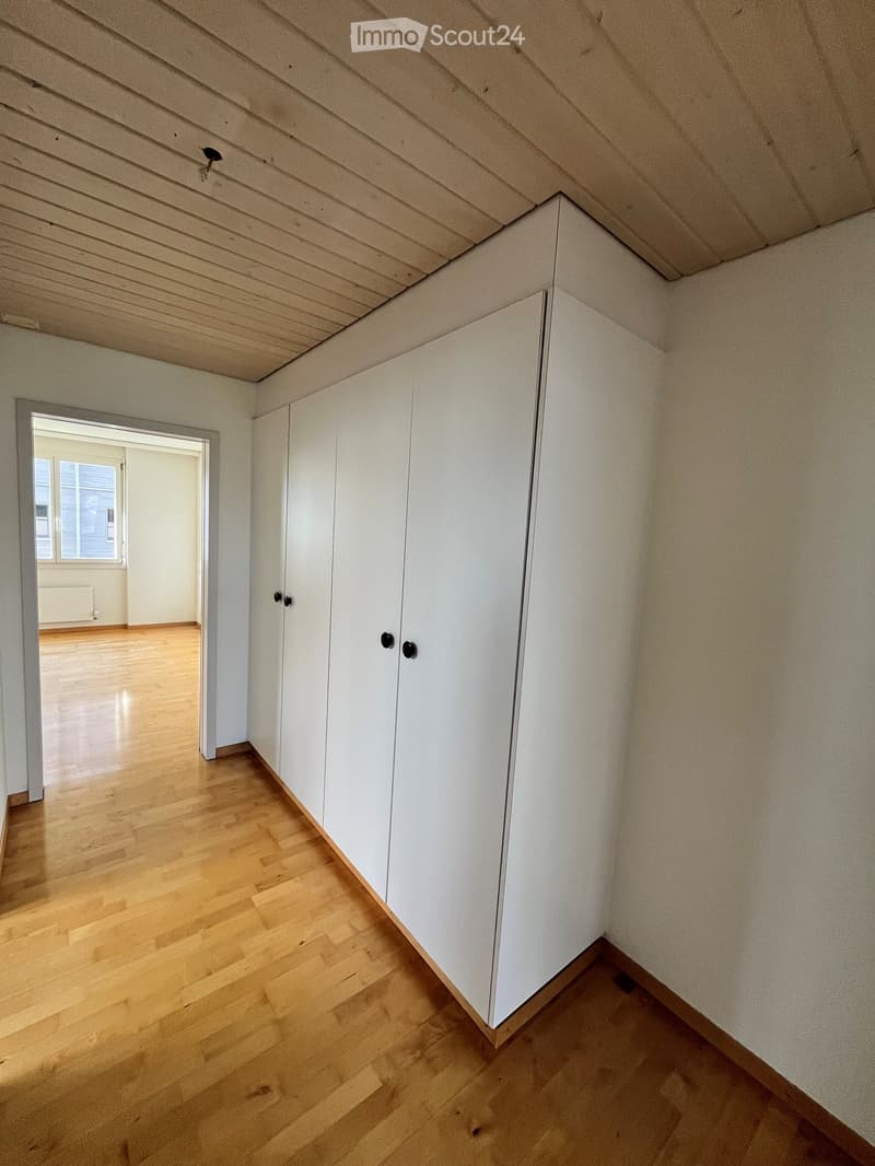 Empty room with light wooden flooring, white walls, and built-in white cabinets. The room has a slanted ceiling and an open doorway leading to another room with a window.