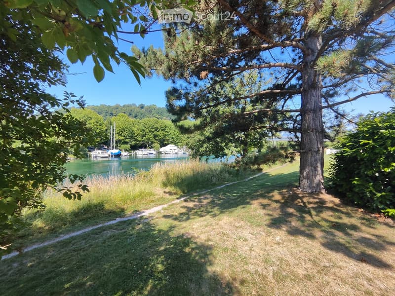 A grassy area with a pathway leading to a waterfront, featuring trees, a body of water, and docked boats in the background.