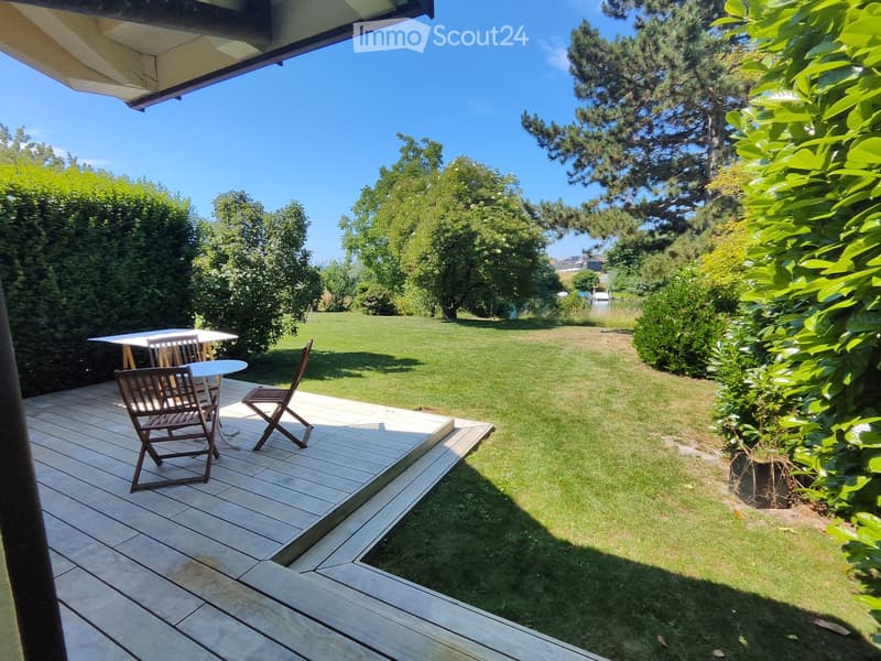 Wooden deck with a table and chairs, overlooking a well-maintained lawn and trees.