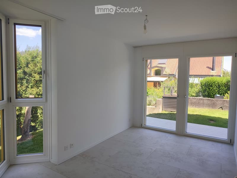 Empty room with white walls, tiled floor, large windows, and sliding glass doors leading to a patio and garden.