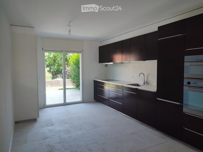 Modern kitchen with dark cabinets, white countertops, and a large window overlooking a garden.