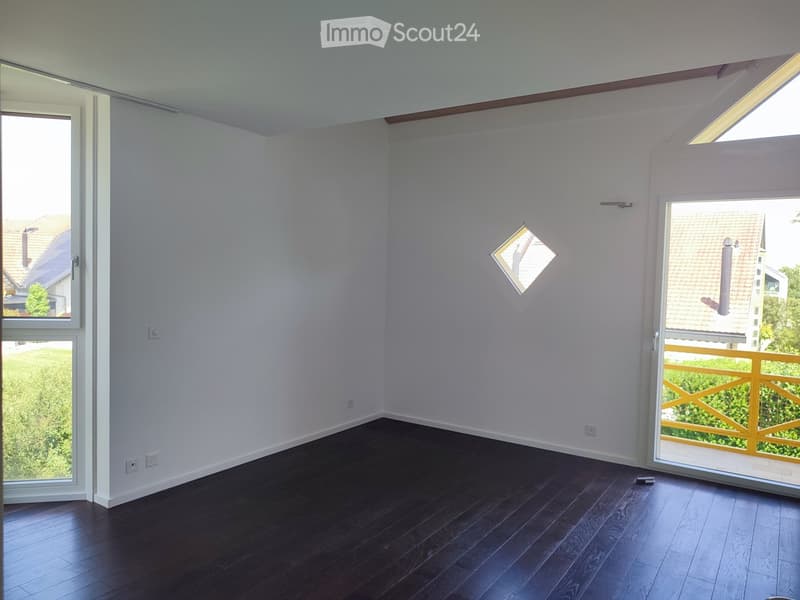 Empty room with dark wooden flooring, white walls, and large windows offering views of the outdoors. The room features a sliding glass door leading to a balcony with a yellow railing.
