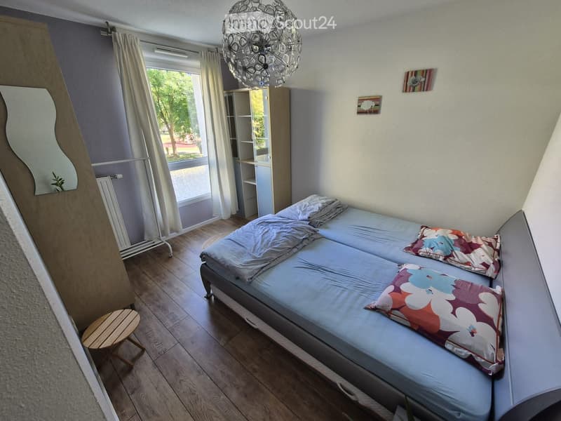 Cozy bedroom with a double bed, two pillows, a small wooden stool, a large mirror, a cabinet, and a decorative chandelier.