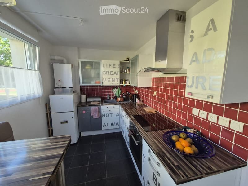 A compact kitchen with white cabinets, red tiled backsplash, and dark countertops. Features include a refrigerator, dishwasher, oven, and a bowl of fruit on the counter.