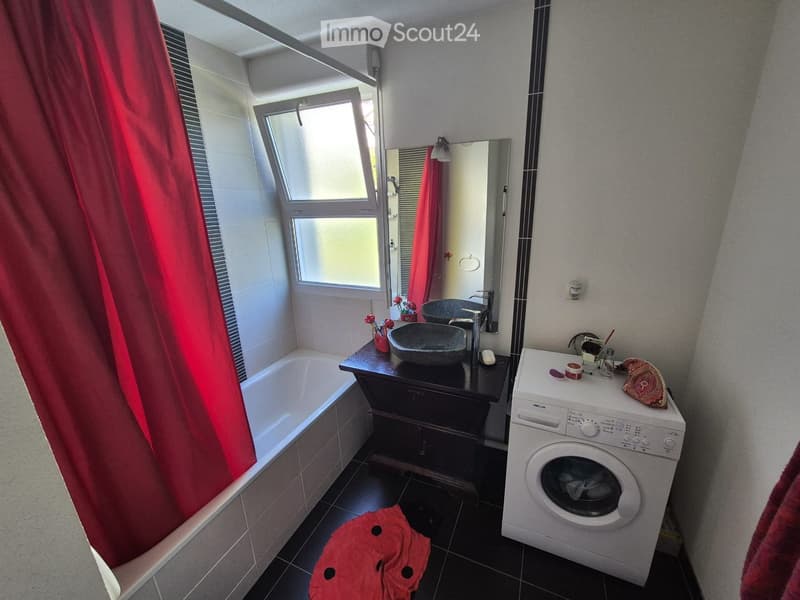 Modern bathroom with a bathtub, sink, mirror, and washing machine. Features a large window with red curtains and a dark tiled floor.