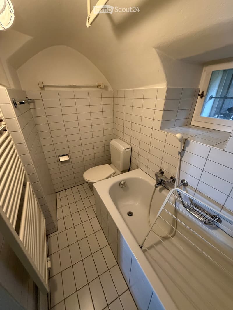 Small bathroom with white tiled walls and floor, featuring a bathtub, toilet, and a radiator. A small window provides natural light, and a shower rack is mounted on the wall.