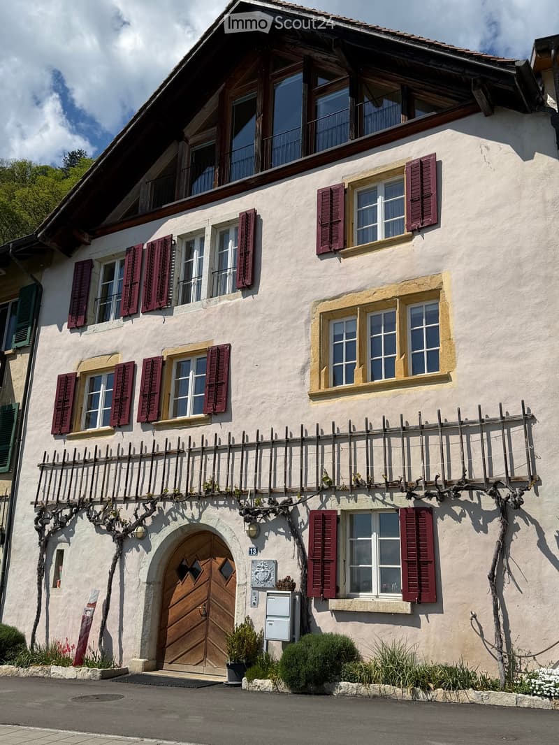 Two-story house with white plaster walls, red shutters, and a wooden balcony with grapevines. Features a large arched wooden door and multiple windows with shutters.
