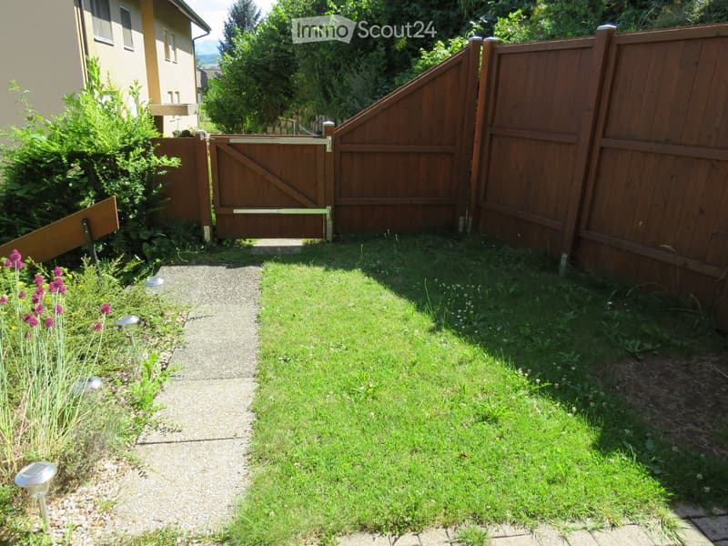A well-maintained garden with a gravel pathway leading to a wooden gate, surrounded by green grass, shrubs, and small purple flowers. The area is enclosed by a wooden fence and features solar-powered garden lights along the pathway.