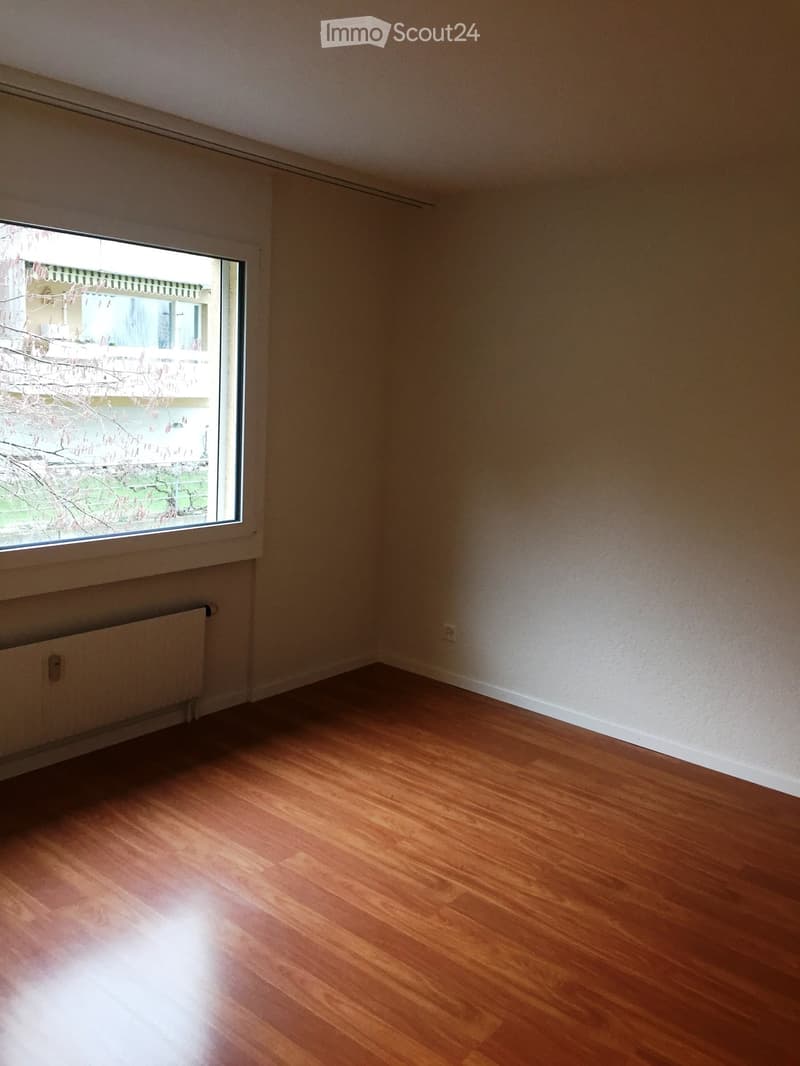 Empty room with wooden flooring, white walls, and a large window with a view of a building and trees outside.