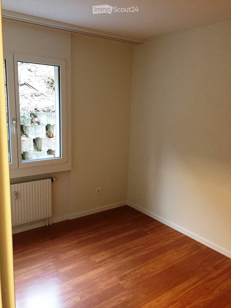 Empty room with wooden flooring, light-colored walls, and a window with a view of a stone wall and greenery outside.