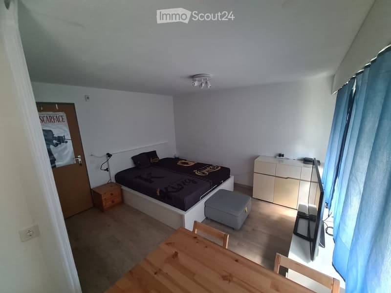 A small, modern bedroom with a single bed, nightstand, dresser, stool, small table, and TV. The room features light-colored walls, wooden flooring, and a large window with blue curtains.