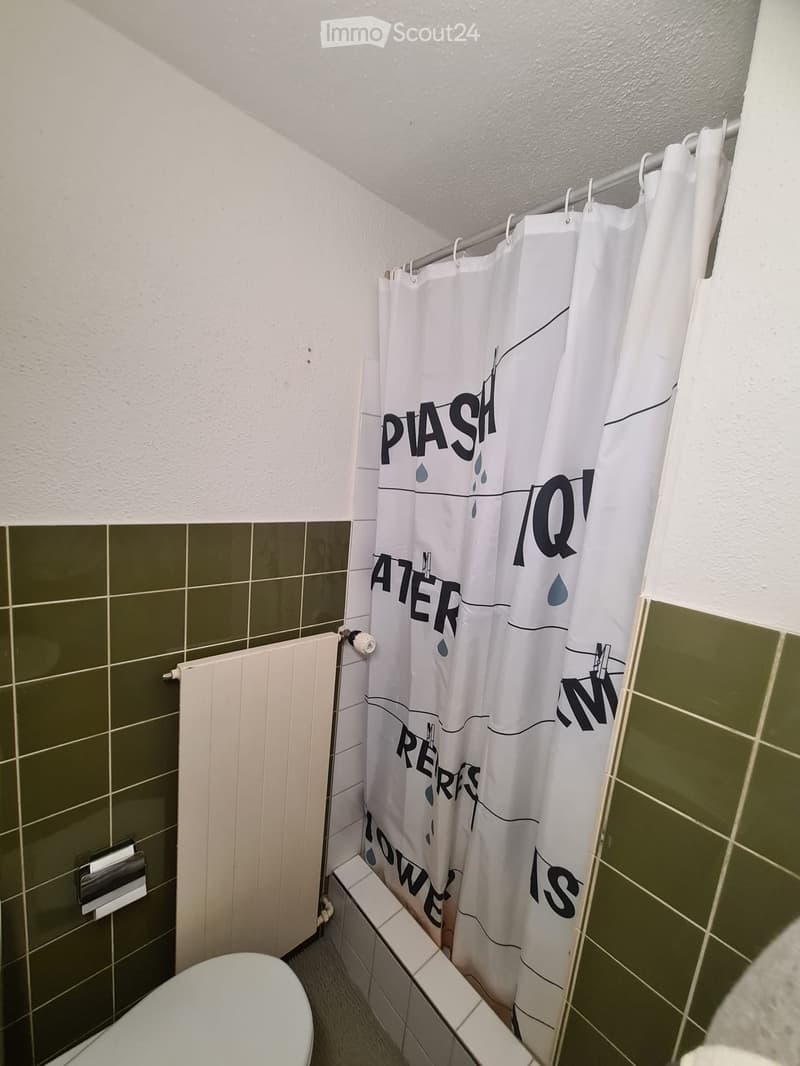 Bathroom with green tiled walls, white shower curtain featuring text, toilet, radiator, and toilet paper holder.