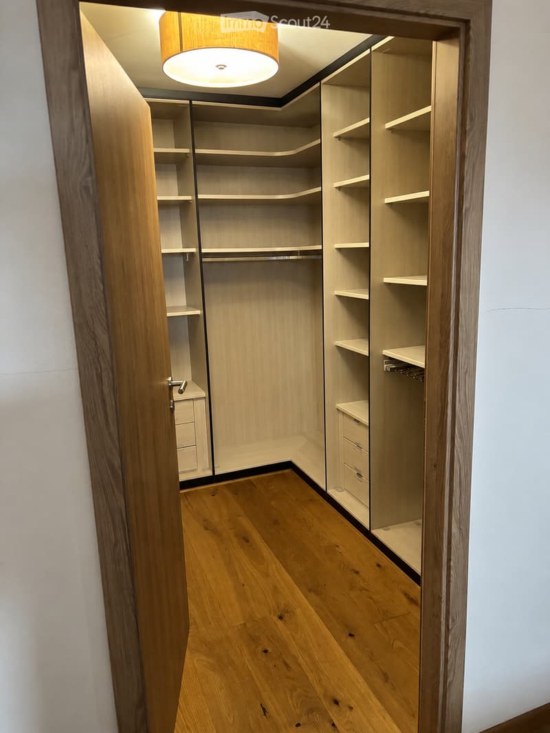 Empty walk-in closet with wooden shelves, drawers, and a hanging rod, featuring light wood finishes and a wooden floor.