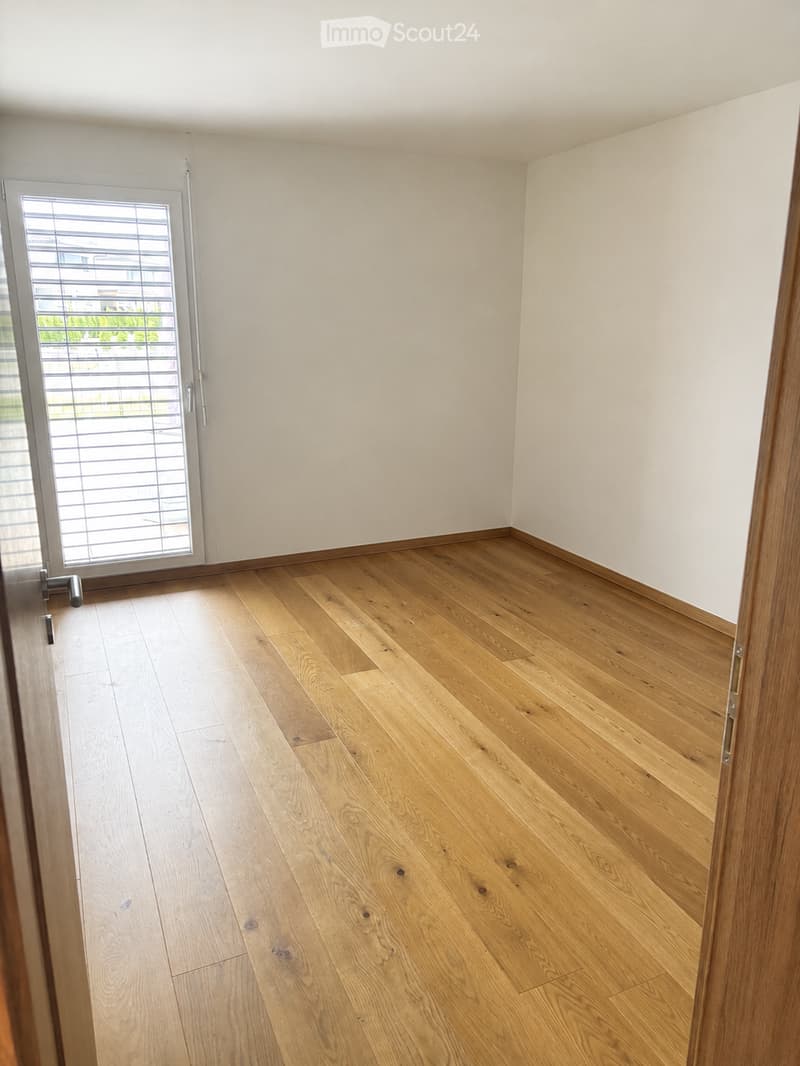 Empty room with light wooden flooring and white walls, featuring a large window with blinds and a door.