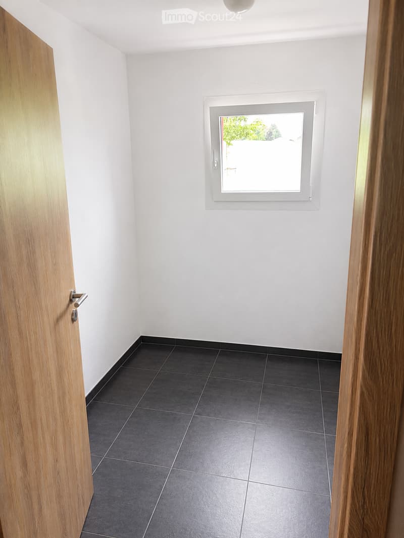 Empty room with white walls, dark grey tiled floor, and a single window with a view of greenery outside.