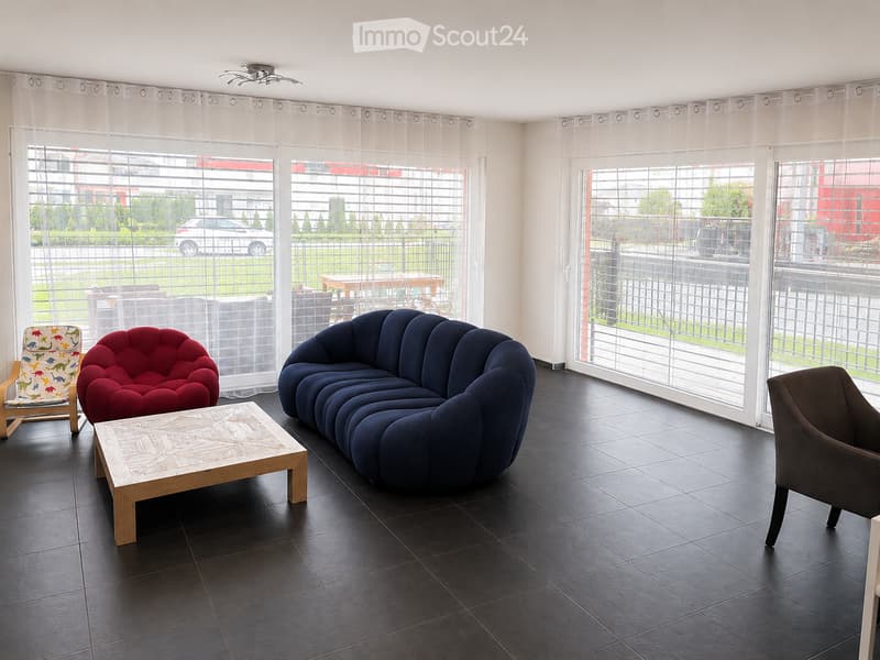 Modern living room with large windows, dark tiled floor, blue sofa, red chair, wooden coffee table, and colorful children's chair.