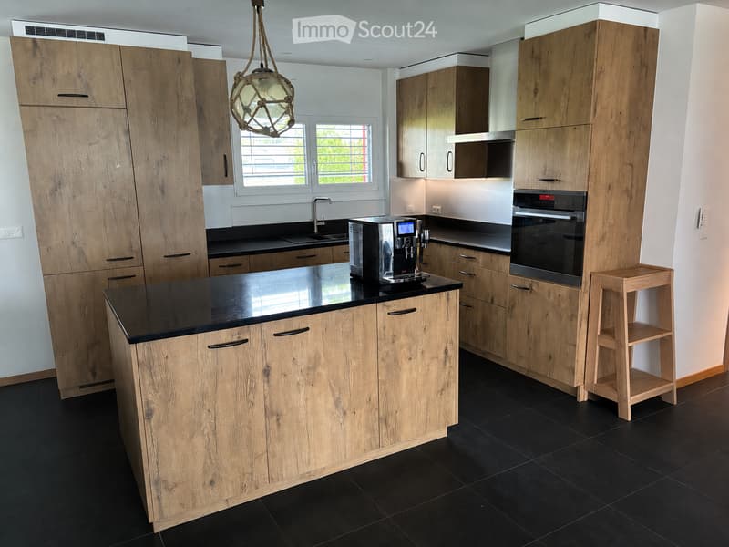 Modern kitchen with wooden cabinets, black countertops, a coffee machine, an oven, and a small wooden stool.