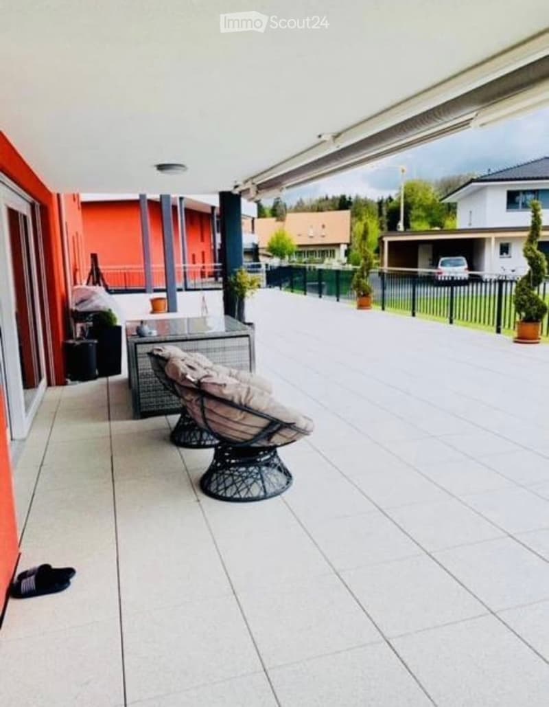 A modern balcony with a tiled floor, a wicker hanging chair, a small table, and potted plants. The balcony is covered with a roof and overlooks a landscaped area with other buildings and greenery.