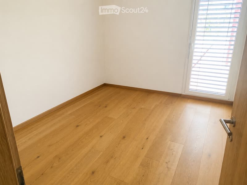 Empty room with light wooden flooring and white walls, featuring a large window with blinds and a door handle visible on the right side.
