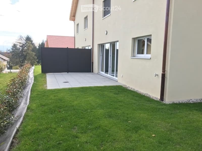 Well-maintained garden area with a neatly trimmed lawn, a paved patio, and a black gate leading to a driveway. The house features large windows and a modern design.