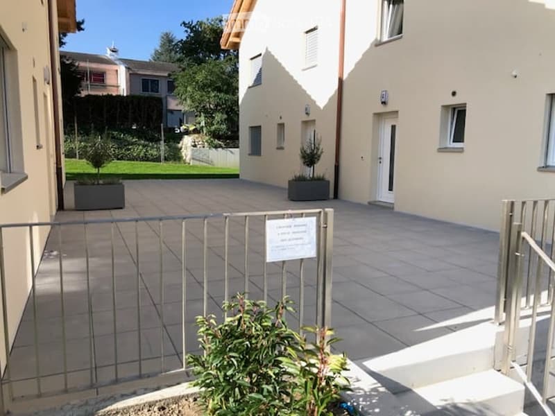 A modern residential complex entrance with a paved courtyard, metal gate, small potted plants, and a neatly trimmed lawn.