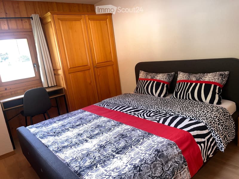 Cozy bedroom with a double bed, patterned bedding, a wooden wardrobe, a desk with a chair, and a window with curtains.