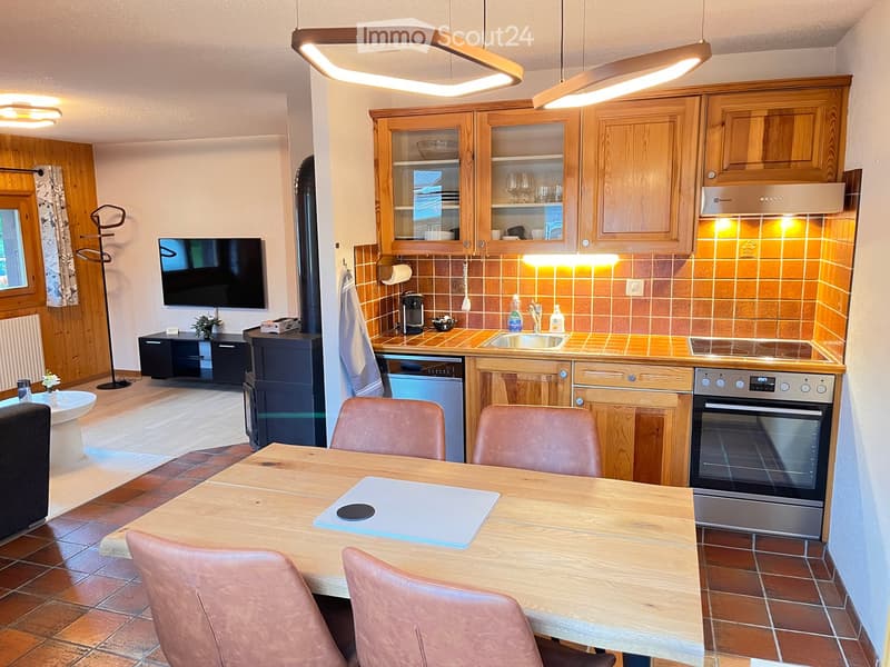 The image shows a cozy, well-furnished room that combines a kitchen and living area. The kitchen features wooden cabinets, a sink, a dishwasher, an oven, and a coffee maker. A wooden dining table with four leather chairs is placed in the center. A televis